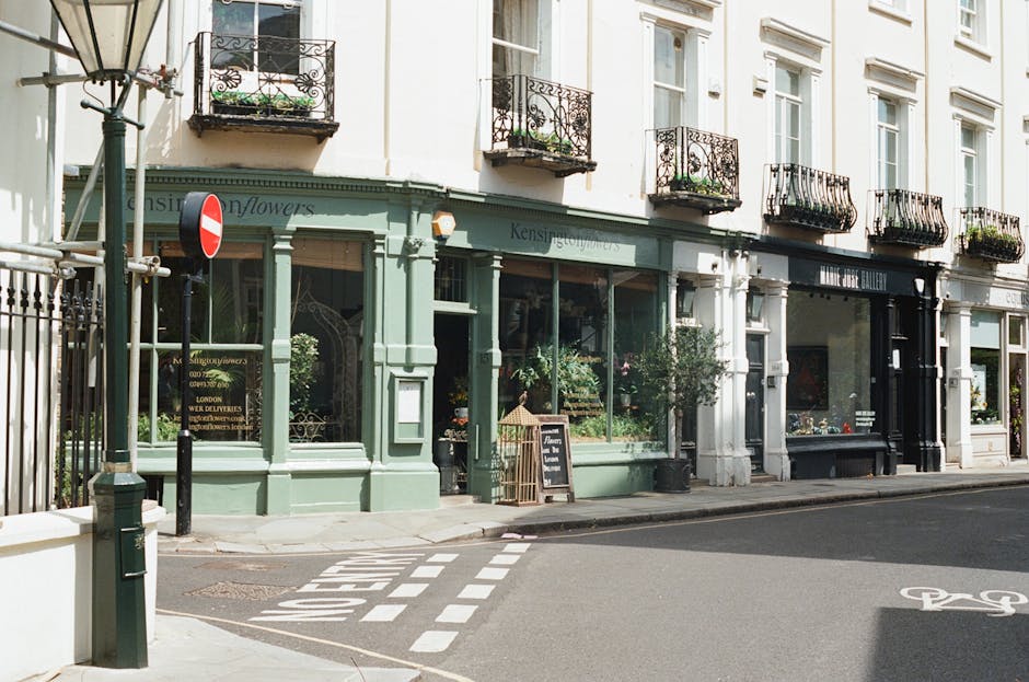 Exterior view of a row of charming storefronts on a city street, featuring a flower shop with a pastel green facade and large display windows filled with vibrant floral arrangements. The shop's window