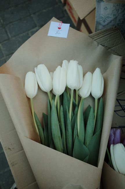 A bouquet of nine white tulips with long green stems and broad leaves, wrapped in light brown kraft paper. The tulips are arranged neatly with their petals closed, and the wrapping is secured with a w