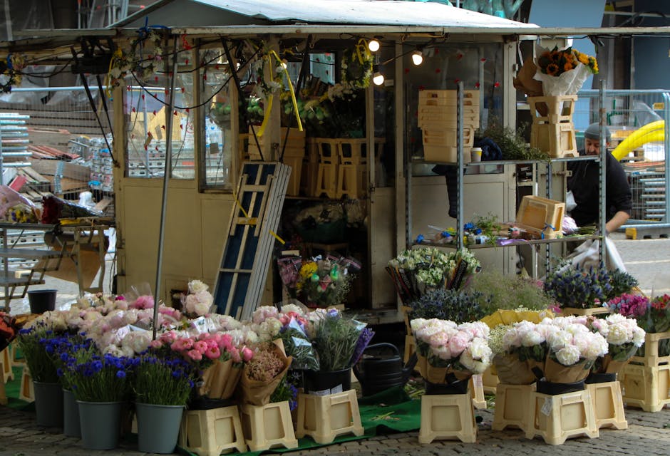 A flower stall displaying a variety of fresh floral bouquets and arrangements on the street, with an assortment of pink, white, purple, and blue flowers including peonies, carnations, and lavender, ar